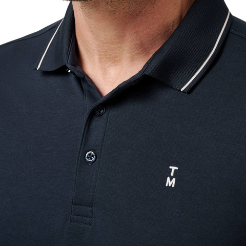 Close-up of a person wearing the Travis Mathew Mens Featherweight Ticket Polo Shirt in Total Eclipse Navy, featuring a collared neckline with white trim, two buttons undone, and small white T M initials embroidered on the left chest.