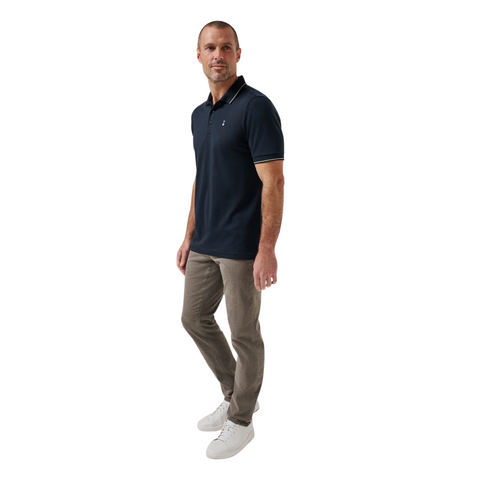 A man stands against a white background wearing the TravisMathew Mens Featherweight Ticket Polo Shirt in Total Eclipse Navy, along with light brown pants and white sneakers. He faces slightly to the side in a relaxed posture.