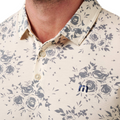 Close-up of a person wearing the TravisMathew Mens Local Forecast Polo Shirt in Birch, featuring a gray floral pattern, blue rh logo on the chest, and buttons for a lightweight, elegant look.