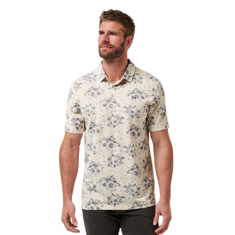 A man with short hair and a beard wears the TravisMathew Mens Local Forecast Polo Shirt in birch—a light, short-sleeve shirt with a subtle floral print—paired with dark pants, standing against a plain white background.