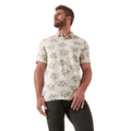 A bearded man wears the TravisMathew Men's Local Forecast Polo Shirt in birch—a cream polo with blue floral pattern—paired with dark pants, standing against a white background and looking left.