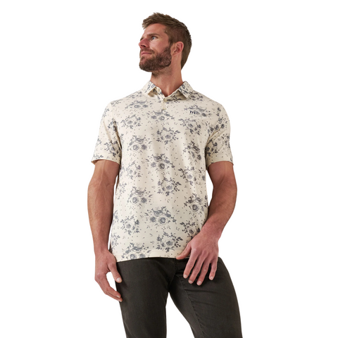 A bearded man wears the TravisMathew Men's Local Forecast Polo Shirt in birch—a cream polo with blue floral pattern—paired with dark pants, standing against a white background and looking left.