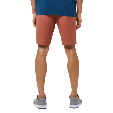 A person is shown from the waist down, facing away, wearing a blue shirt, gray sneakers, and Travis Mathew Mens Step Dance 9 Shorts in Maple against a white background.