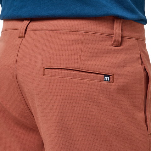 Close-up of a person wearing rust-colored Travis Mathew Mens Step Dance 9 Shorts in Maple with a textured pattern, paired with a blue shirt. These lightweight stretch shorts have a back pocket with a small logo patch above it.