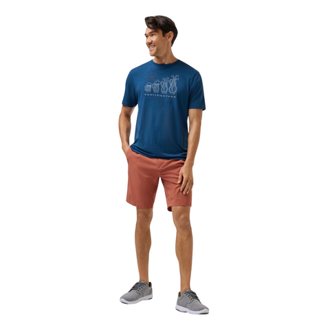 A man smiles with his hands in his pockets, wearing a blue graphic tee, TravisMathew Mens Step Dance 9 Shorts in Maple by Travis Mathew, and gray sneakers as he looks to the side against a white background.