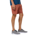 A person wearing a blue shirt, Travis Mathew Men's Step Dance 9 Shorts in Maple, and gray athletic shoes stands facing left, shown from mid-torso to feet against a white background.