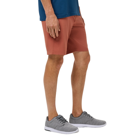 A person wearing a blue shirt, Travis Mathew Men's Step Dance 9 Shorts in Maple, and gray athletic shoes stands facing left, shown from mid-torso to feet against a white background.