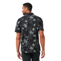 A man faces away, wearing the Travis Mathew Trek Tide Mens Polo—a black, short-sleeve button-up with a gray floral pattern and moisture-wicking fabric—paired with black pants against a white background.