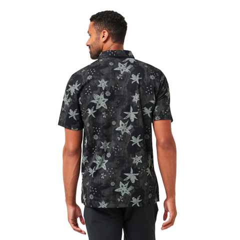 A man faces away, wearing the Travis Mathew Trek Tide Mens Polo—a black, short-sleeve button-up with a gray floral pattern and moisture-wicking fabric—paired with black pants against a white background.