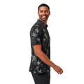 A man with short hair and a beard smiles while walking in a moisture-wicking, dark Travis Mathew Trek Tide Mens Polo with a bold floral print and dark pants against a plain white background.