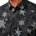 Close-up of a person wearing the Travis Mathew Mens Trek Tide Polo, featuring a dark floral pattern, moisture-wicking fabric, button placket, and a small branded logo on the chest.