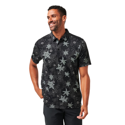 A man smiles in the Travis Mathew Trek Tide Mens Polo—a moisture-wicking, short-sleeve gray floral golf shirt—paired with dark pants, set against a white background.