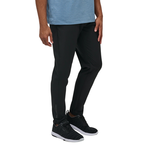 A man stands with arms relaxed, wearing the TravisMathew Open To Close Men’s Jogger in black by Travis Mathew, paired with a blue shirt and black sneakers, highlighting the joggers all-day comfort from shoulders down.