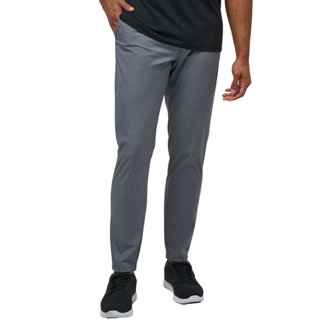 A man stands against a white background wearing Travis Mathew Open To Close Men's Jogger pants in gray, paired with a black shirt and black sneakers.