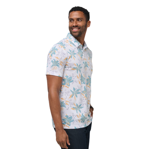 A man with short hair and a beard smiles to his left, wearing dark pants and a blue-and-yellow floral Travis Mathew Paradise Peak Men’s Polo, displaying stylish modern men’s golf apparel against a plain background.