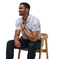 A man with short curly hair, wearing a Travis Mathew Paradise Peak Men’s Polo in white and blue patterns and dark jeans, sits sideways on a wooden chair, smiling as he adjusts his collar with one hand.