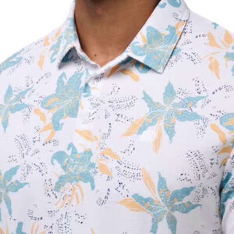 A close-up of a person wearing the Travis Mathew Paradise Peak Men’s Polo, a white golf shirt with blue and yellow floral designs. The image highlights the collar, buttons, and upper chest—ideal men’s golf apparel from Travis Mathew.