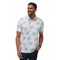 A man wearing the Travis Mathew Paradise Peak Men’s Polo, a short-sleeve golf shirt with a blue, green, and yellow floral design, stands smiling with one hand in his pocket and a watch on his wrist.