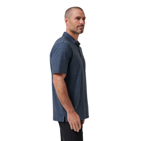 A man with a trimmed beard and short hair stands in profile, wearing the Travis Mathew The Heater Mens Polo—a breathable performance polo—paired with black pants against a plain white background.