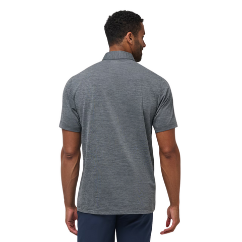 A man stands facing away, showing the back of the moisture-wicking TravisMathew The Heater Pro Mens Polo in heather gray, paired with navy pants, against a plain white background.