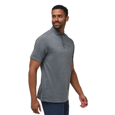 A man in a Travis Mathew The Heater Pro Mens Polo—a short-sleeved, gray, moisture-wicking shirt—paired with dark pants stands sideways, looking over his shoulder with a neutral expression against a white background.