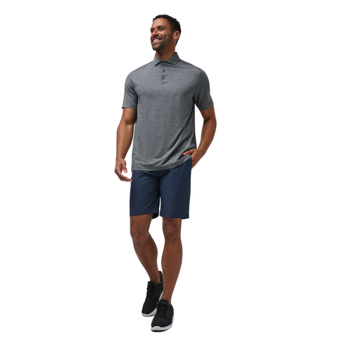 A man wearing a Travis Mathew The Heater Pro Mens Polo stands smiling with one hand in his pocket, paired with navy shorts and black athletic shoes against a white background.