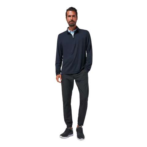 A man with short dark hair and a beard stands against a white background wearing the TravisMathew The Heater Quarter Zip Mens Long Sleeve Polo, dark joggers, a light blue shirt underneath, and black athletic shoes, with one hand in his pocket.