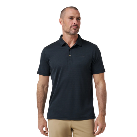 A man with short hair and a beard wears a black TravisMathew The Zinna 2.0 Men’s Polo and beige pants, standing against a plain white background.