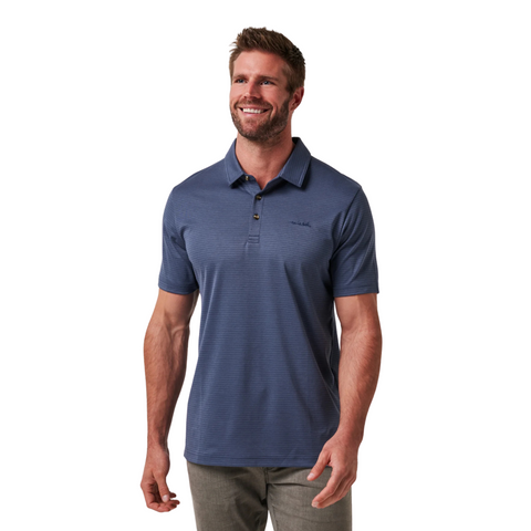 A man with short brown hair and a beard smiles, wearing the Travis Mathew The Zinna 2.0 Men’s Polo in Mood Indigo and gray pants, standing against a white background.