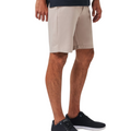 A man models beige Travis Mathew Wanderlust Mens Shorts with a dark shirt and black athletic shoes, standing with hands at his sides, shown from mid-torso to feet against a plain white background.