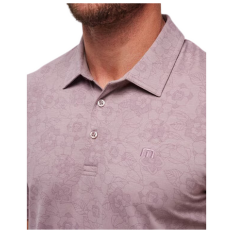 Close-up of the TravisMathew New Destination Polo on a model, highlighting the soft floral pattern on the muted purple fabric, three-button placket, and embroidered TravisMathew logo on the chest. The texture adds subtle detail to the classic design.