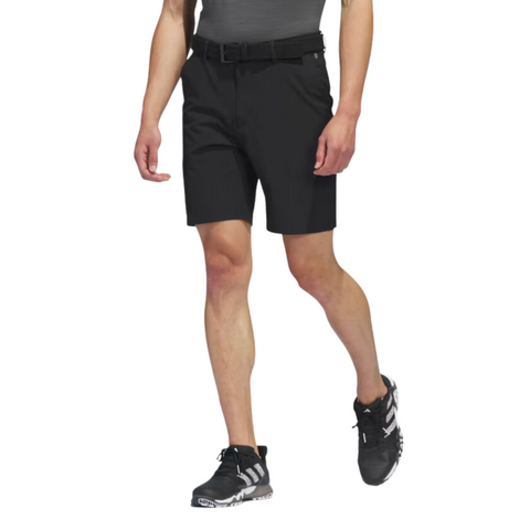 The Adidas Ultimate365 8.5-Inch Men's Golf Shorts in black, worn by a model, feature a tailored fit with front pockets and a belt, paired with black Adidas golf shoes.