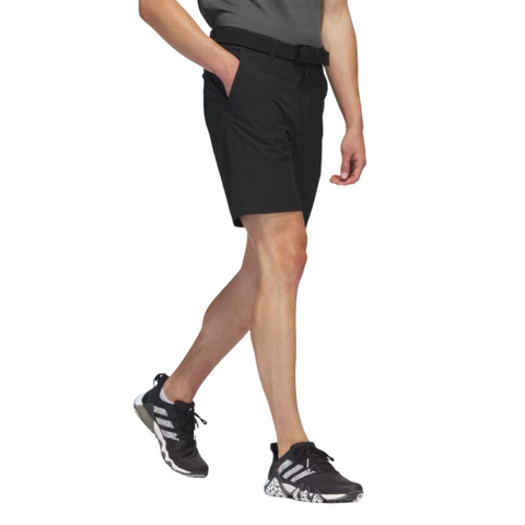 The Adidas Ultimate365 8.5-Inch Men's Golf Shorts in black, worn by a model, feature a tailored fit with side pockets and a belt, paired with black Adidas golf shoes.