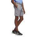 The Adidas Ultimate365 8.5-Inch Men's Golf Shorts in grey, worn by a model, side view, features a tailored fit with side pockets and a belt, paired with black Adidas golf shoes.